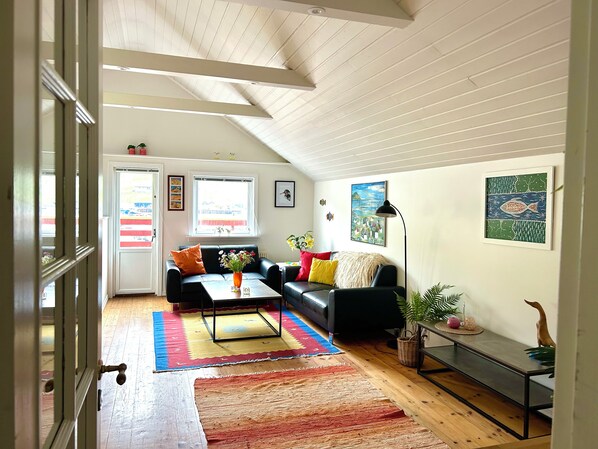 A living room with a black sofa, a coffee table, and a TV. There are two windows with curtains, a painting on the wall, and a door leading to another room.