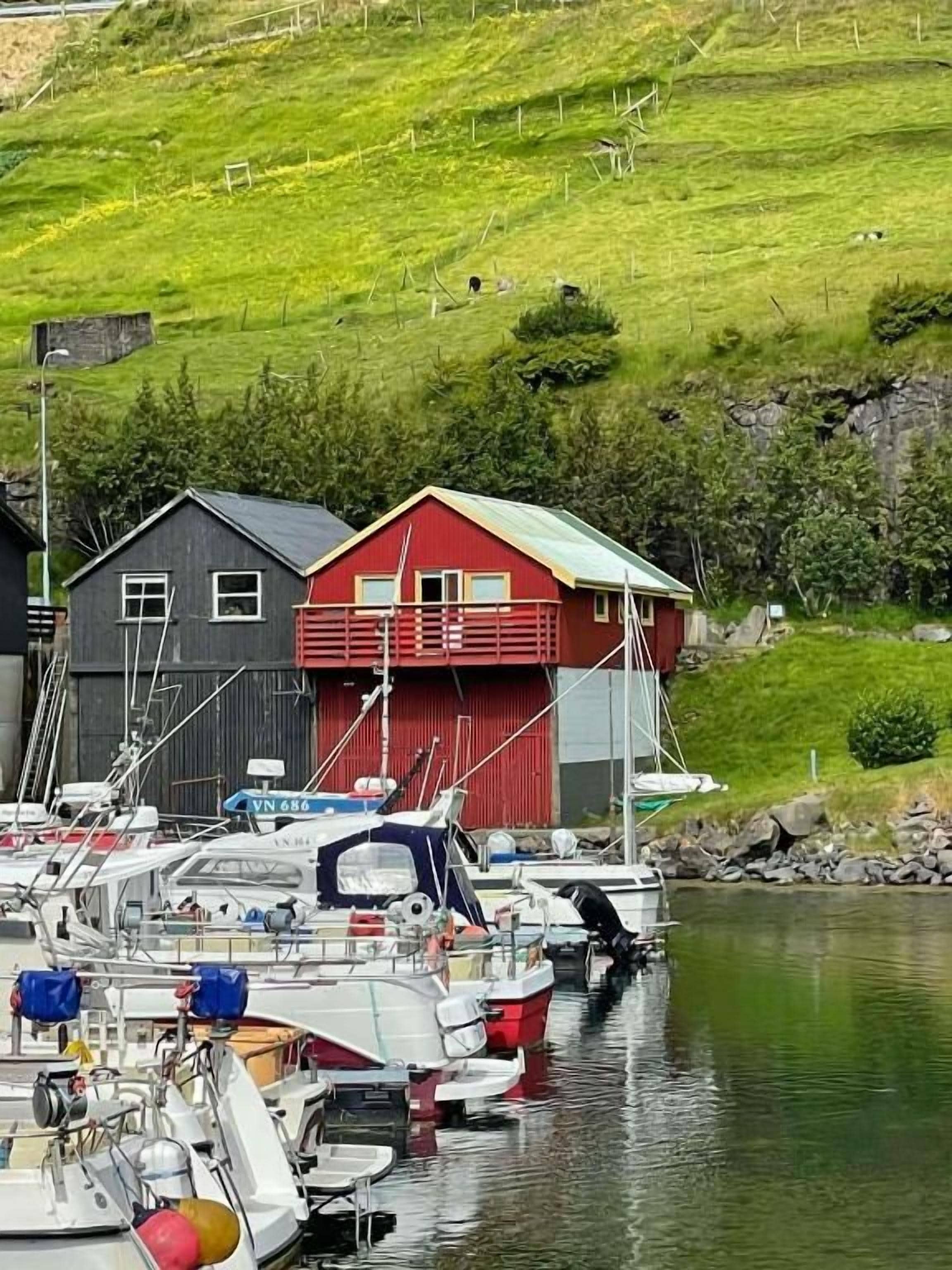Ein Yachthafen mit vertäuten Booten, ein rotes Gebäude und ein grasbewachsener Hügel im Hintergrund.