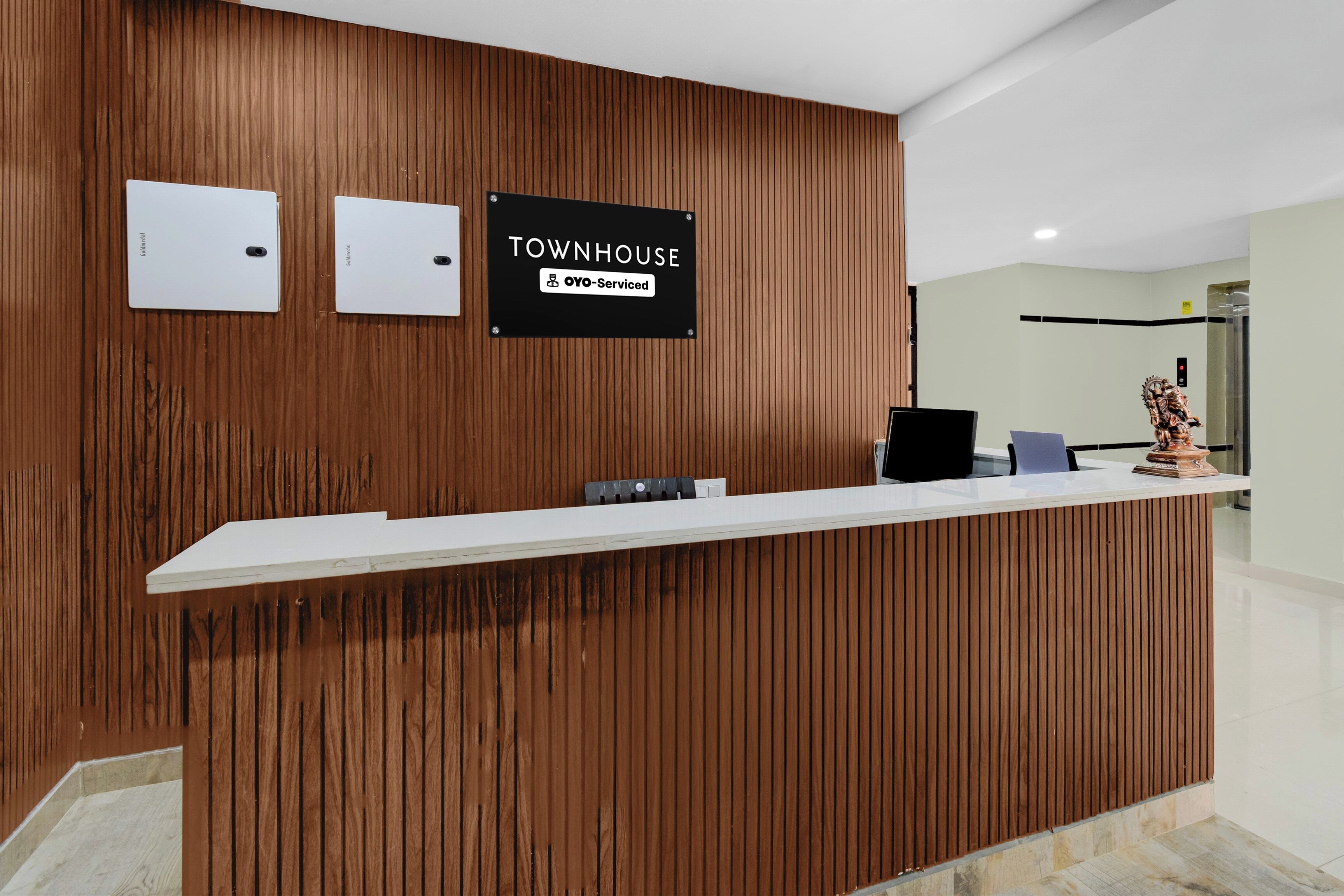 Un mostrador de recepción con un panel de pared de madera, un cartel que indica 'TOWNHOUSE OYO-Serviced', y una zona de recepción con una computadora y una pequeña estatua.