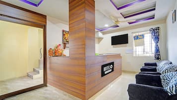 A hotel reception area with a wooden reception desk, a flat-screen TV mounted on the wall, a calendar, and a window with curtains.