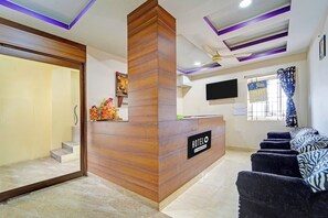 A hotel reception area with a wooden reception desk, a flat-screen TV mounted on the wall, a calendar, and a window with curtains.