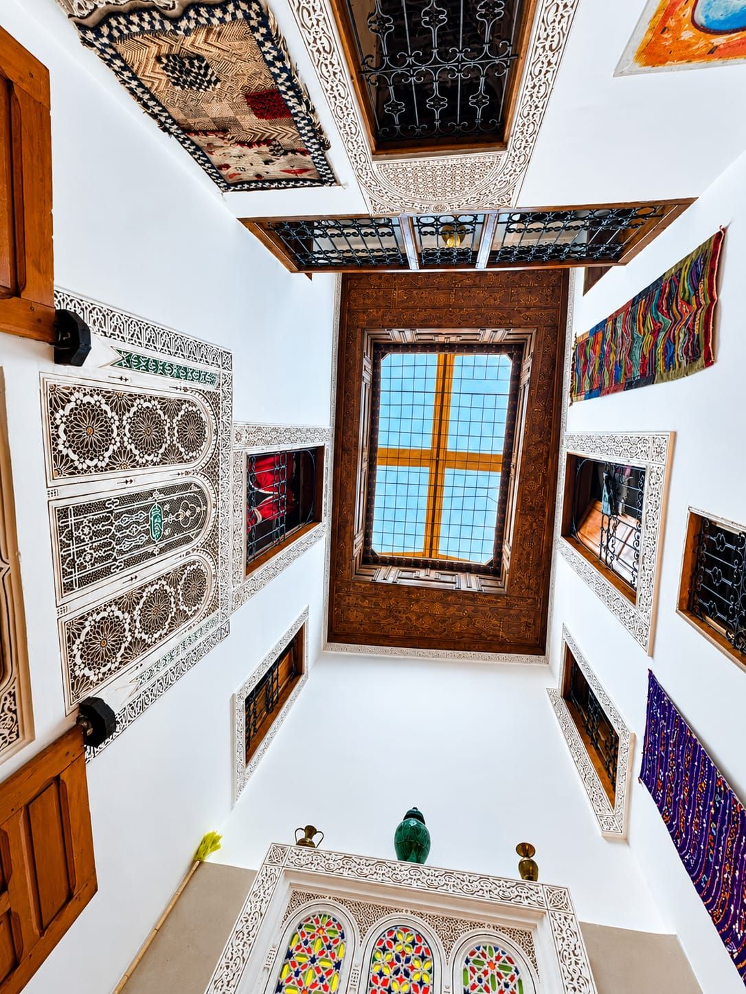 A room with intricate wall and ceiling designs, a central window, and a small table with a green bottle.