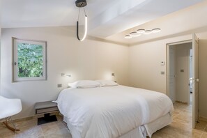 Ein modernes Schlafzimmer mit einem großen Bett, Nachttischlampen, einem Beistelltisch und Blick auf Grün hinter einem Fenster.