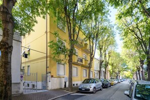 A tree-lined street with parked cars and a yellow building. - GuestHost - Ondina Apartment in Downtown Pesaro (Pesaro)