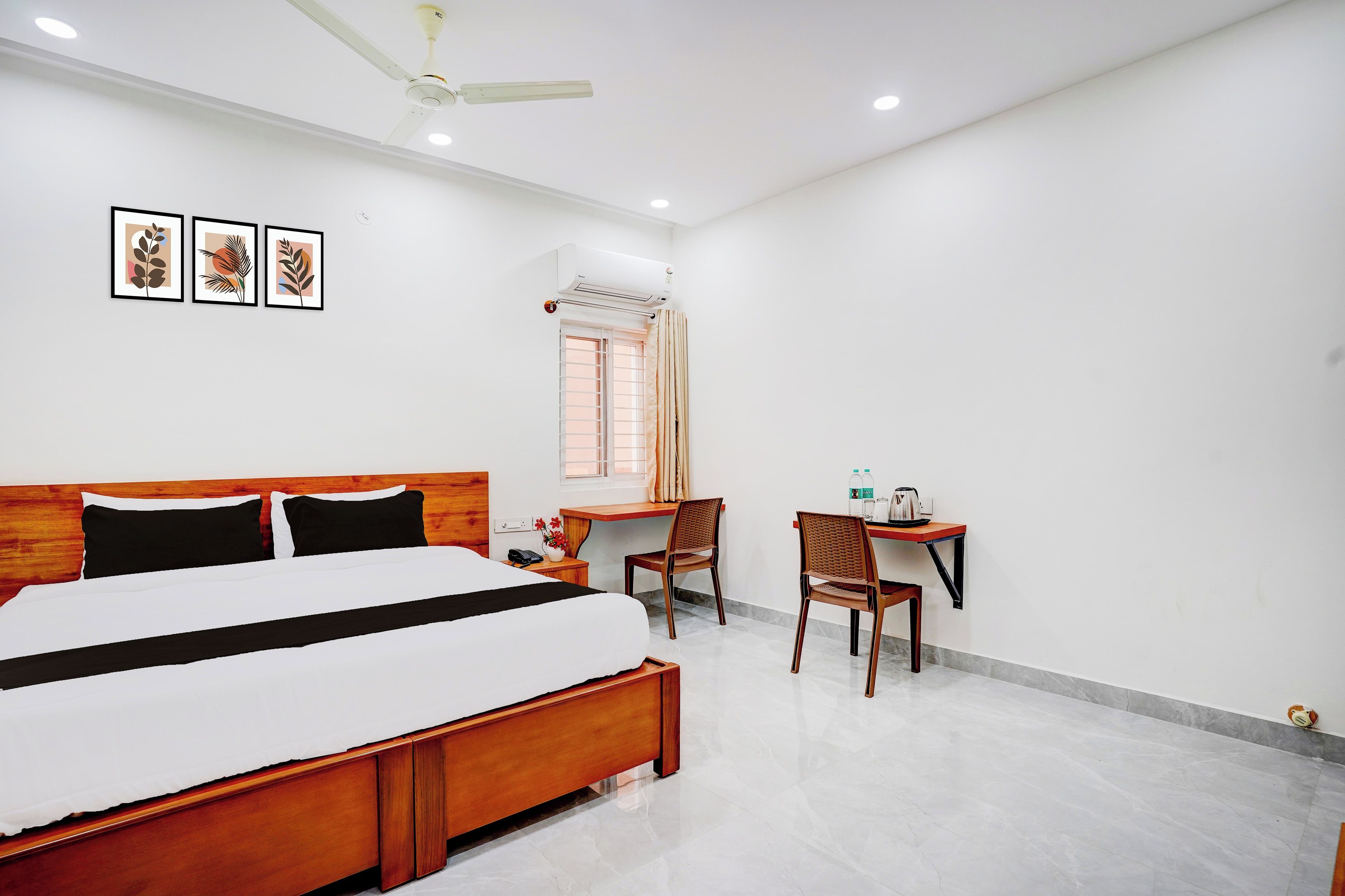A hotel room with a large bed, a desk, two chairs, a window with curtains, and a ceiling fan.