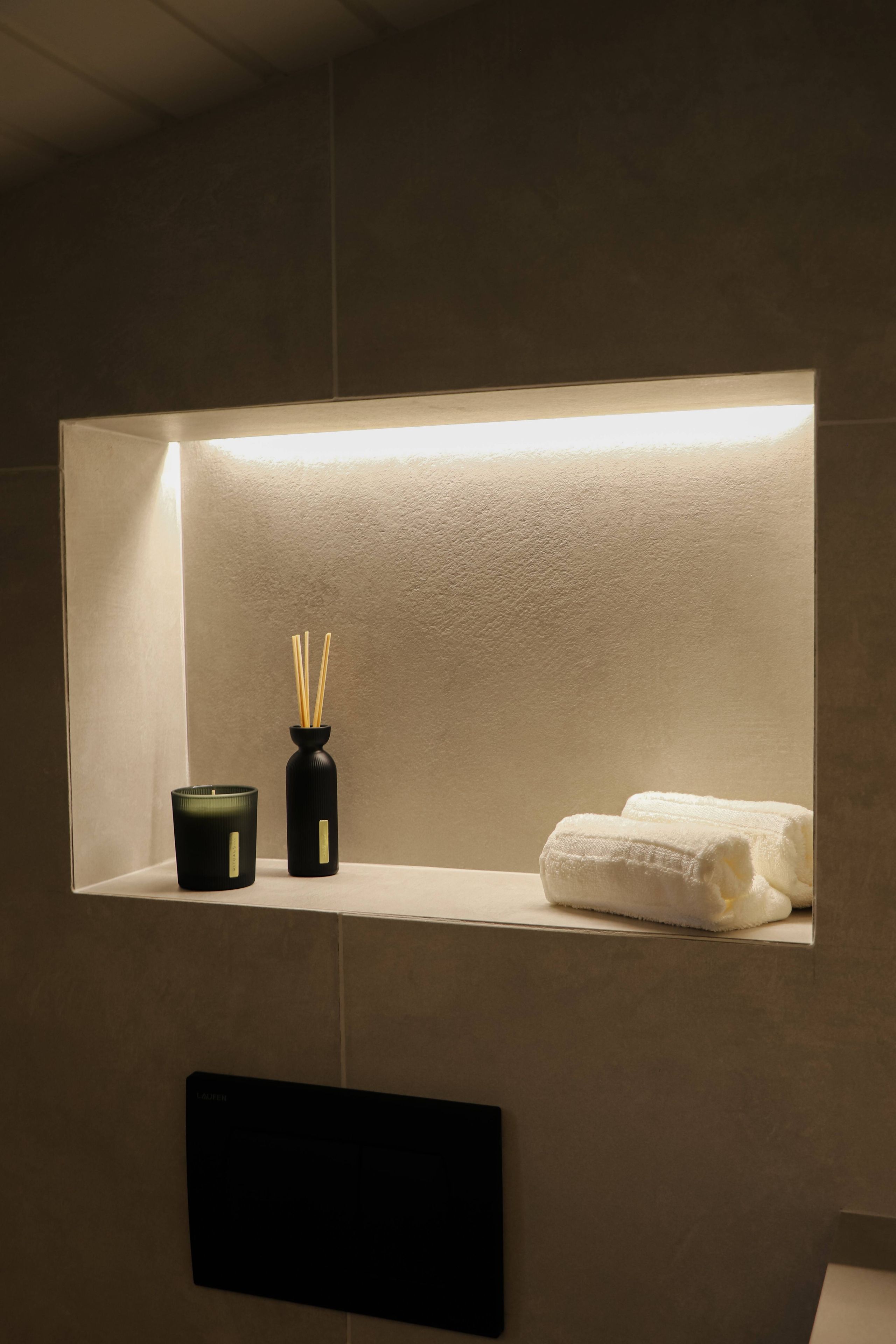 A bathroom with a shelf containing a candle, a vase with reed sticks, and a folded towel.