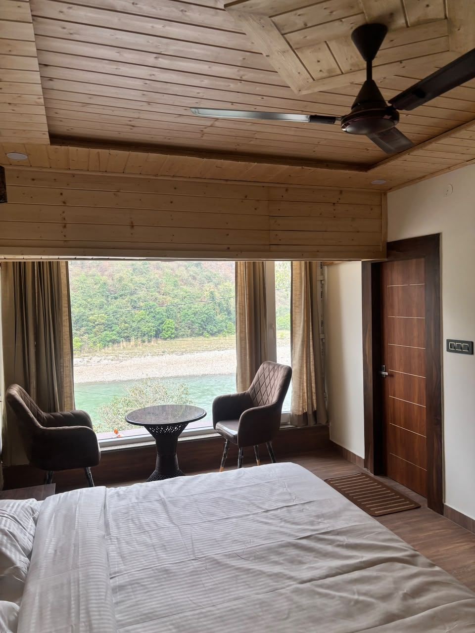 A hotel room with a bed, a ceiling fan, a view of a river, and two chairs by a window.