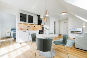 Eine moderne Küche mit weißen Schränken, ein Essbereich mit einem runden Tisch und Stühlen sowie ein Wohnzimmer mit einer Couch und einem Fernseher.