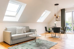 Ein modernes Wohnzimmer mit einem grauen Sofa, einem runden Couchtisch und einem Essbereich mit Tisch und Stühlen. Im Deckenbereich befinden sich Oberlichter und durch die Fenster ist ein Blick ins Freie möglich.