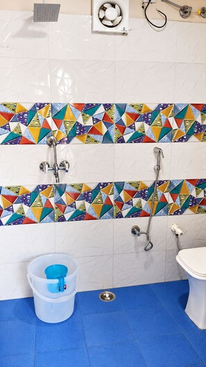A bathroom with a white tiled wall, a colorful geometric tile border, a blue floor, a white toilet, and a shower area with a handheld showerhead. - The Aurum Resort (Mohan Chatti, Silogi Road)