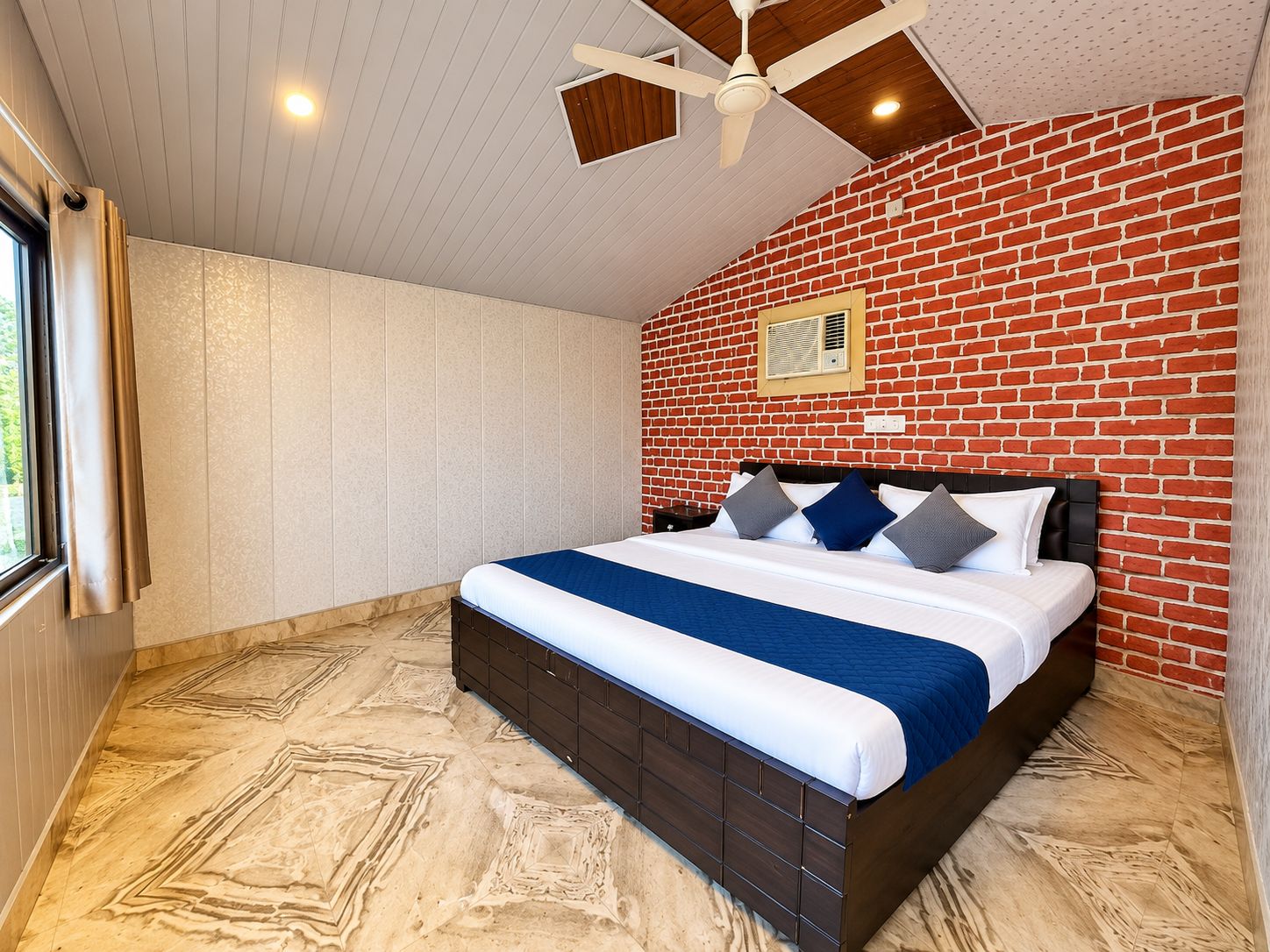 Un dormitorio con pared de ladrillo, cama con colcha azul y blanca, ventilador de techo y ventana con cortinas.