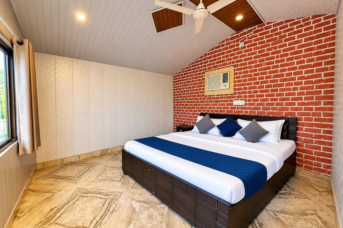 A bedroom with a brick wall, a bed with a blue and white bedspread, a ceiling fan, and a window with curtains.