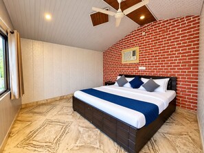 A bedroom with a brick wall, a bed with a blue and white bedspread, a ceiling fan, and a window with curtains. - The Aurum Resort (Mohan Chatti, Silogi Road)