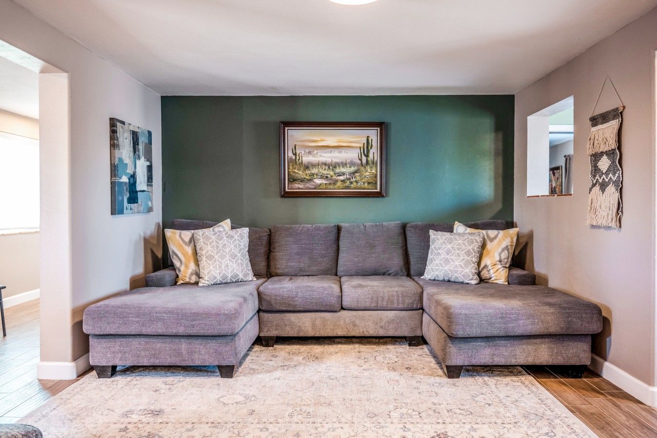 A modern living room with a sectional sofa, a framed landscape painting, and a decorative wall hanging.