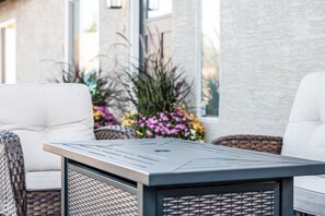 A patio area with a metal table and wicker chairs, surrounded by a building with large windows and a flower arrangement.