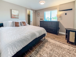 A bedroom with a bed, a dresser, a window with blinds, and a wall-mounted shelf.