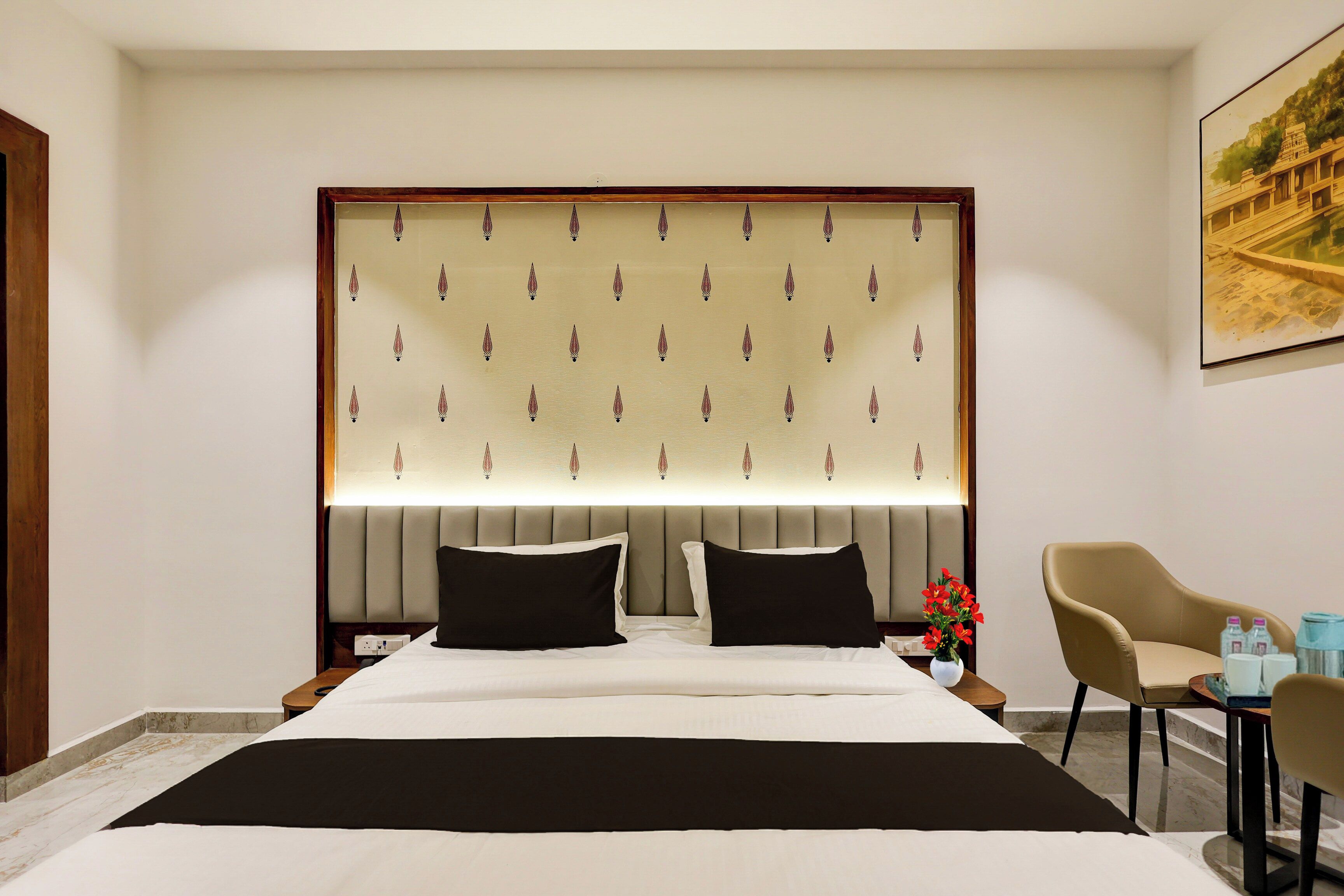 A hotel room with a large bed, a headboard with a patterned design, two bedside tables, a chair, and a painting on the wall.