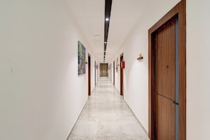 Un long couloir étroit aux murs blancs et au sol en marbre. Des portes se trouvent de part et d’autre, et le couloir est bien éclairé grâce à des luminaires au plafond.