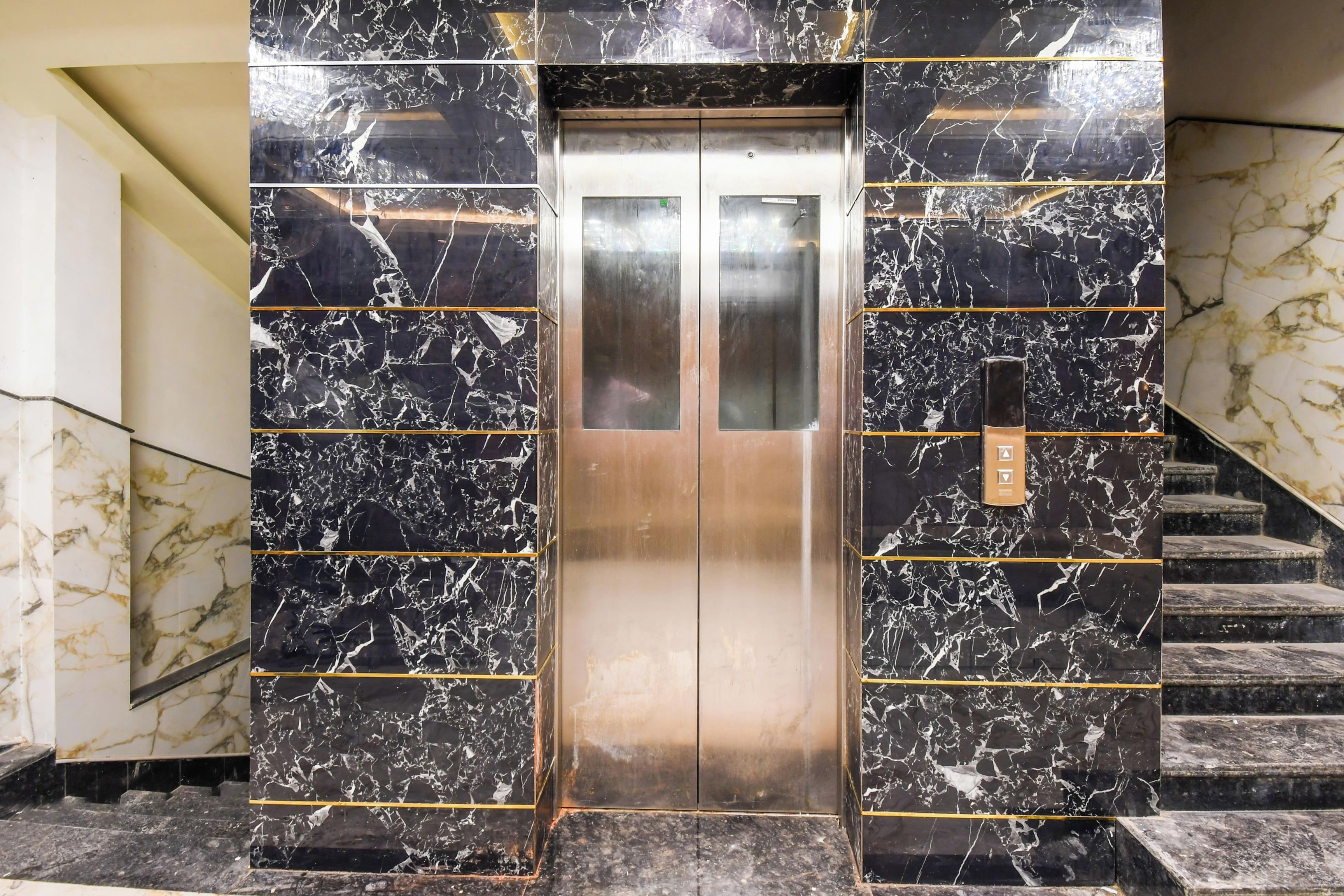 Een lift met een metalen interieur, omringd door marmeren muren met gouden accenten, die uitkomt bij een trap.