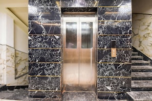 An elevator with a metallic interior, flanked by marble walls with gold accents, leading to a staircase. - Hotel O Krishna Regency (Krishnanagar)