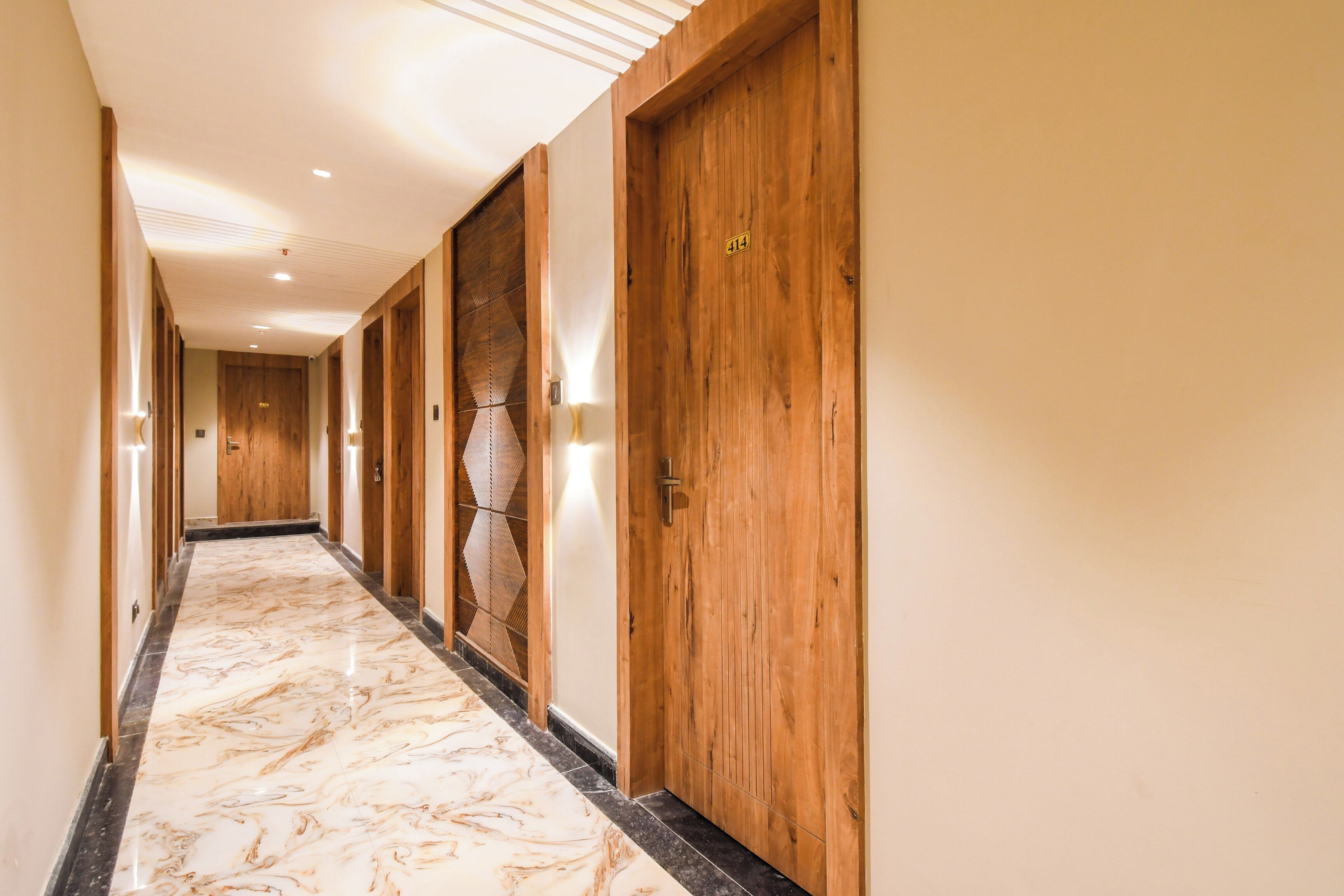 A hotel corridor with wooden doors, marble flooring, and wall-mounted lighting.