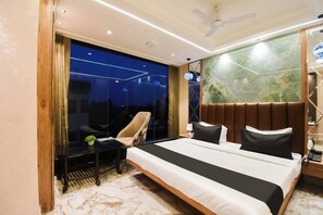 Ein modernes Hotelzimmer mit einem großen Bett, einer Sitzecke inklusive Sessel, einem kleinen Tisch und Blick durch eine Glaswand.