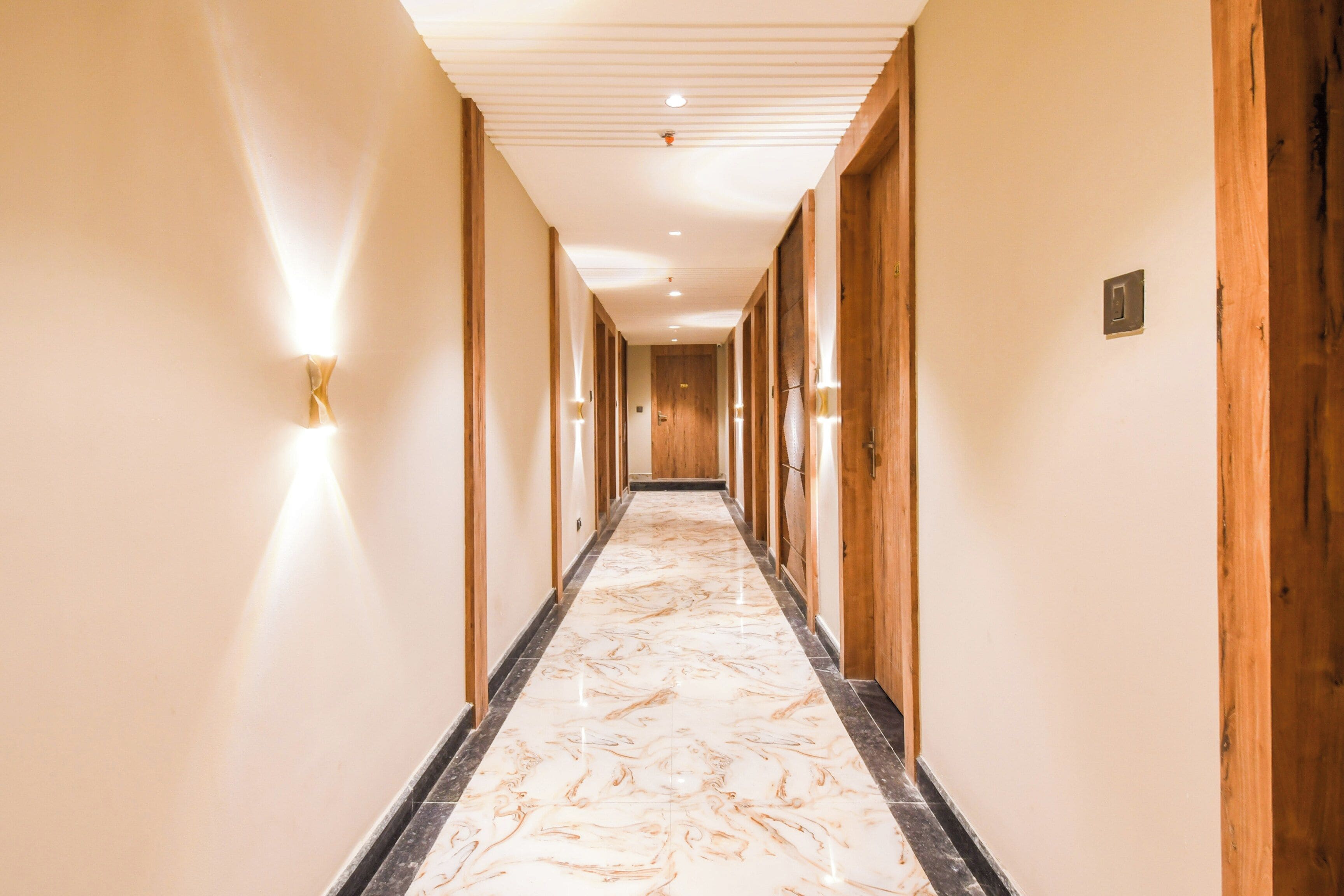 A hotel hallway with marble flooring, wooden doors, and wall-mounted light fixtures.
