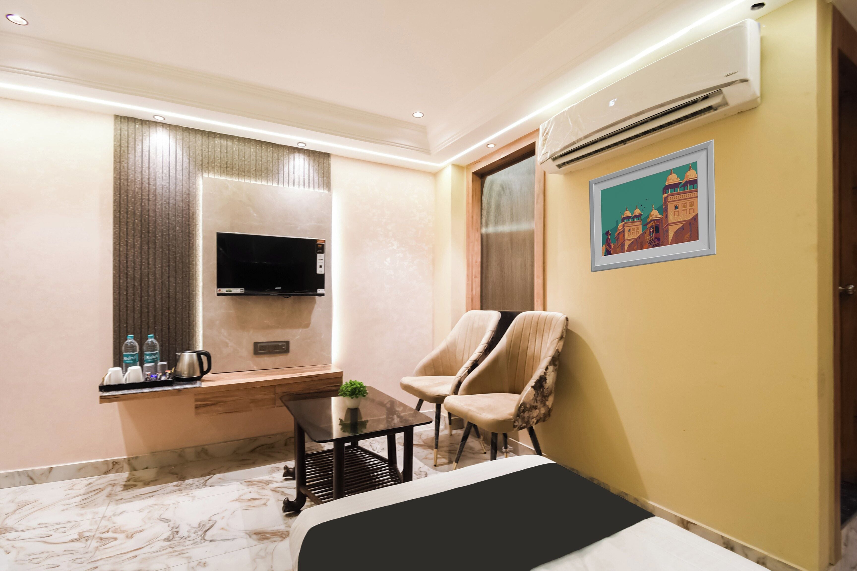 A hotel room with a bed, a TV, a desk with a chair, a small table with a plant, and an air conditioning unit.