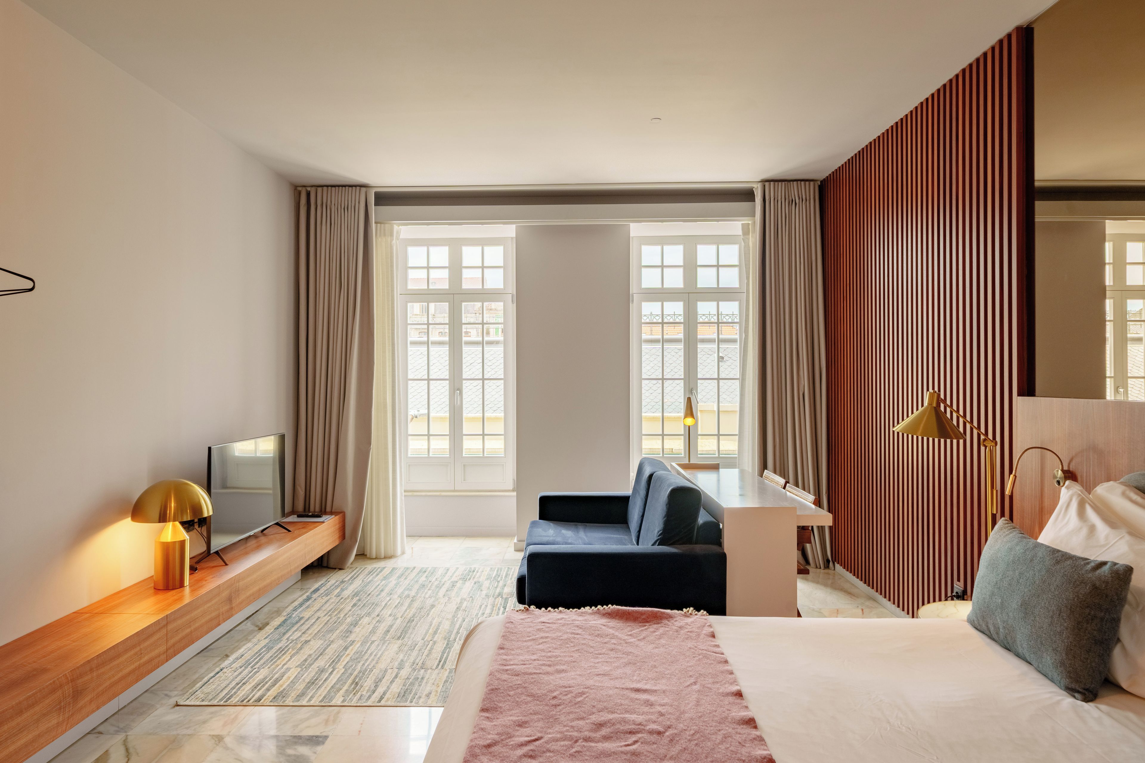 A modern hotel room with a bed, a desk, a sofa, a TV, and large windows.