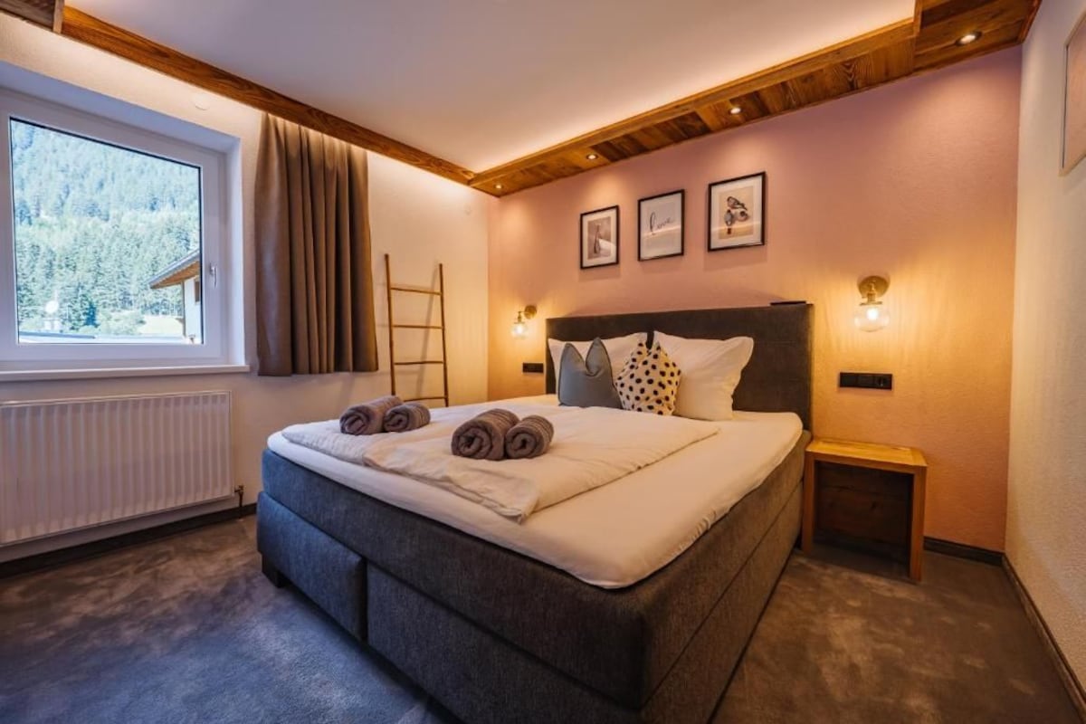 A bedroom with a large bed, a window with curtains, a wooden ceiling, and wall-mounted lights.