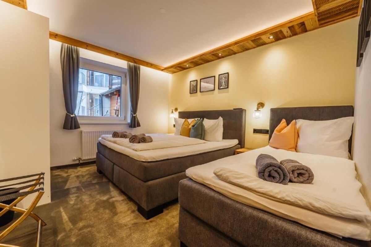 A hotel room with two beds, a window with curtains, a radiator, and a wooden ceiling.