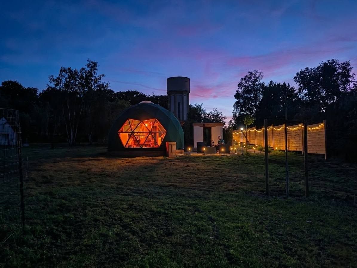 Une structure en forme de dôme géodésique, dotée d’un éclairage intérieur chaleureux, entourée d’un champ d’herbe et d’arbres.