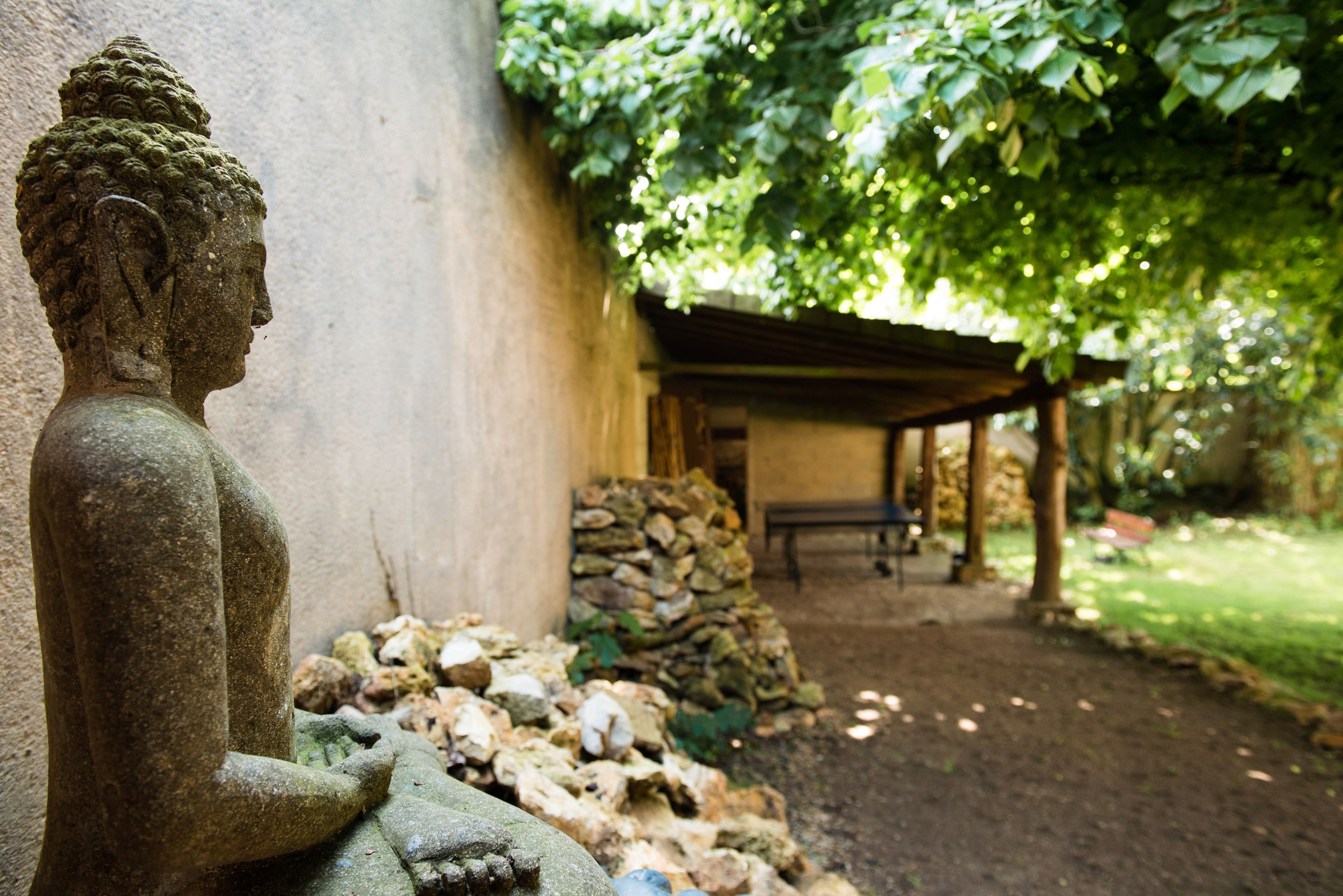 Een stenen Boeddhabeeld in een tuin met een stenen muur, een overdekte zitplaats en een stapel stenen.