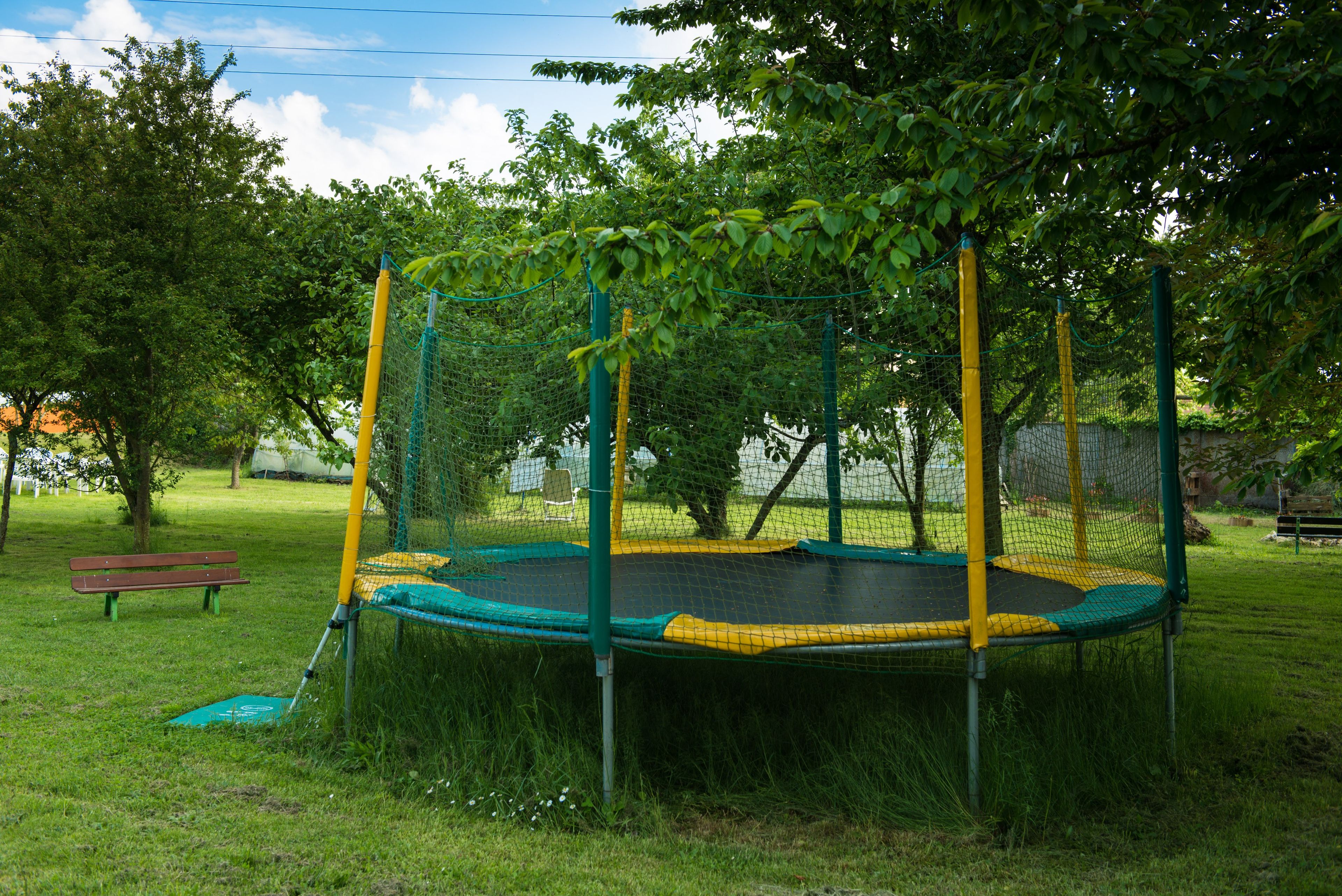 Een trampoline omringd door een veiligheidsnet, gelegen in een grasrijke omgeving met bomen en een bankje op de achtergrond.