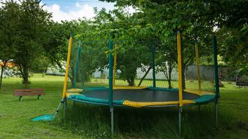 Een trampoline omringd door een veiligheidsnet, gelegen in een grasrijke omgeving met bomen en een bankje op de achtergrond.