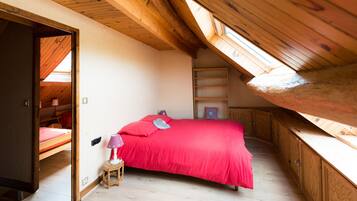 Una camera da letto con soffitto spiovente, un letto con copriletto rosso, uno sgabello di legno e un comodino con una lampada.