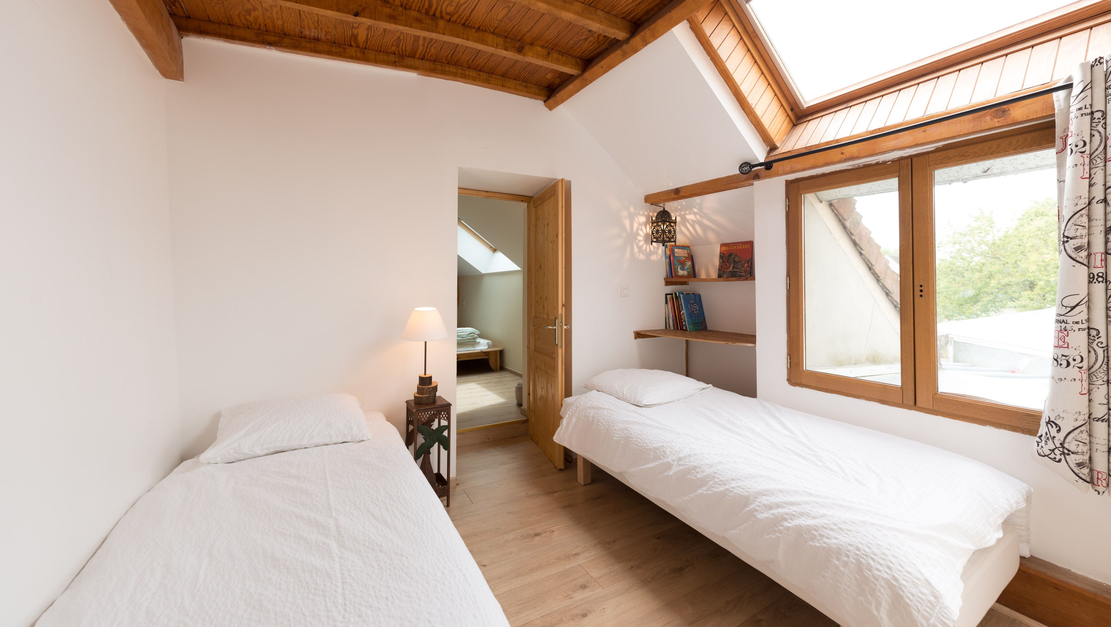 Ein Schlafzimmer mit zwei Betten, einem Oberlicht, einem Fenster mit Vorhängen, einer Nachttischlampe und einem Bücherregal.