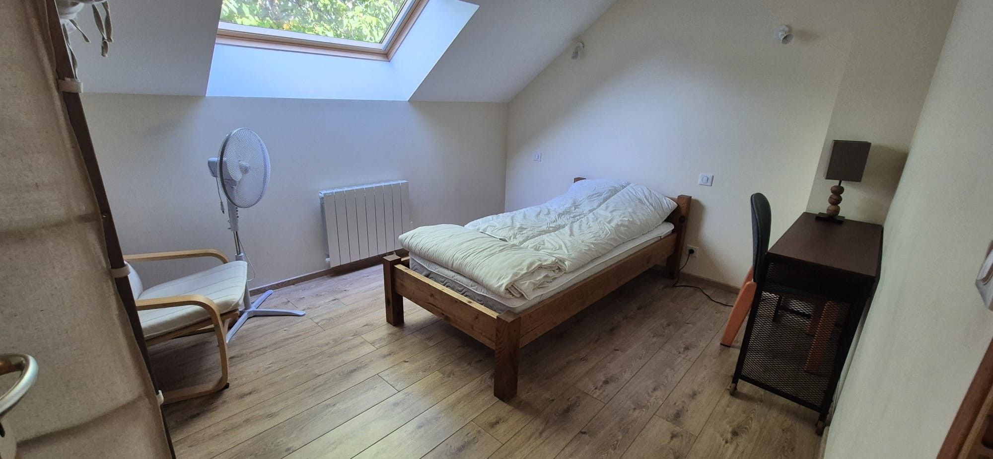 Ein kleines Zimmer mit einem Einzelbett, einer schrägen Decke, einem Heizkörper, einem Stuhl, einem Schreibtisch und einem Fenster.
