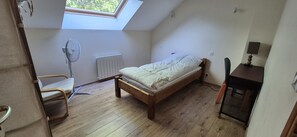 Ein kleines Zimmer mit einem Einzelbett, einer schrägen Decke, einem Heizkörper, einem Stuhl, einem Schreibtisch und einem Fenster.