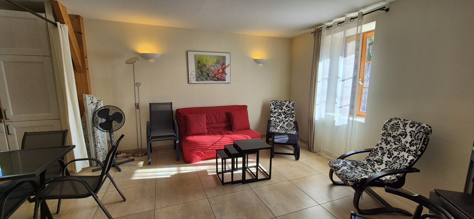 Een woonkamer met een rode bank, twee gestreepte stoelen, een klein tafeltje, een ventilator en een schilderij aan de muur.