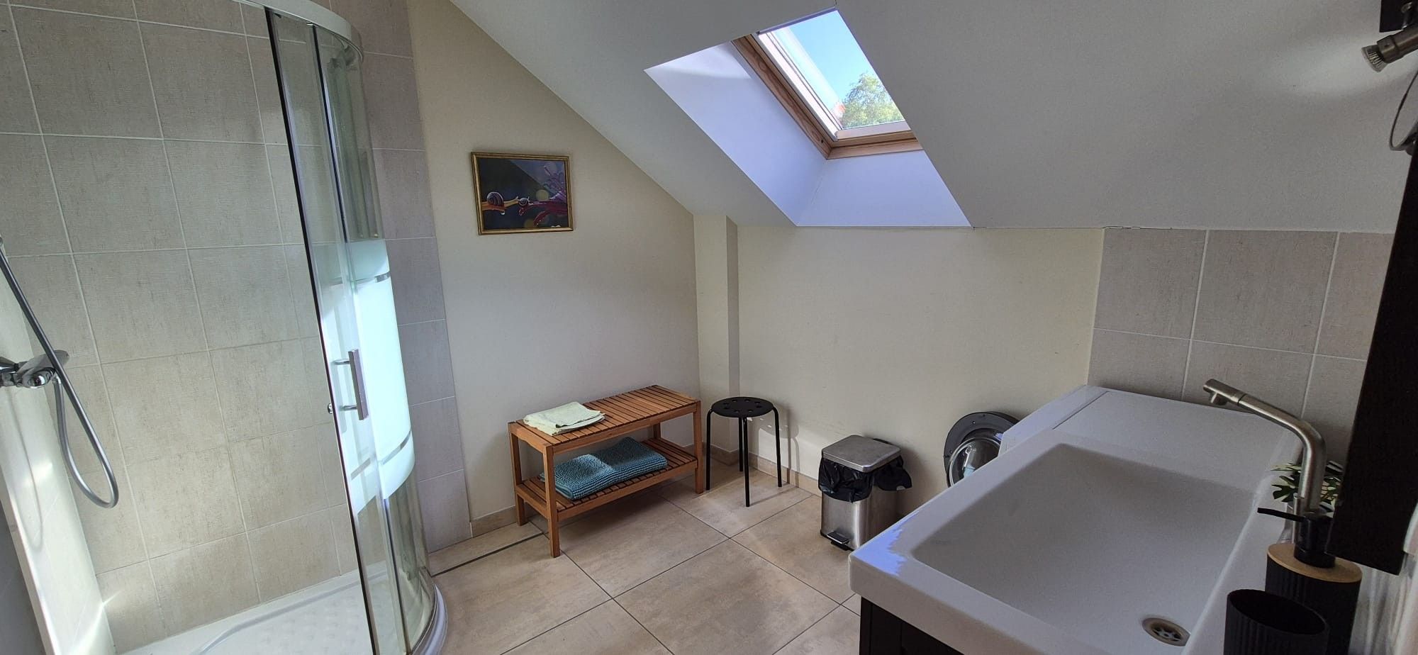 A modern bathroom with a skylight, a glass shower enclosure, a wooden bench, a small stool, and a large bathtub.