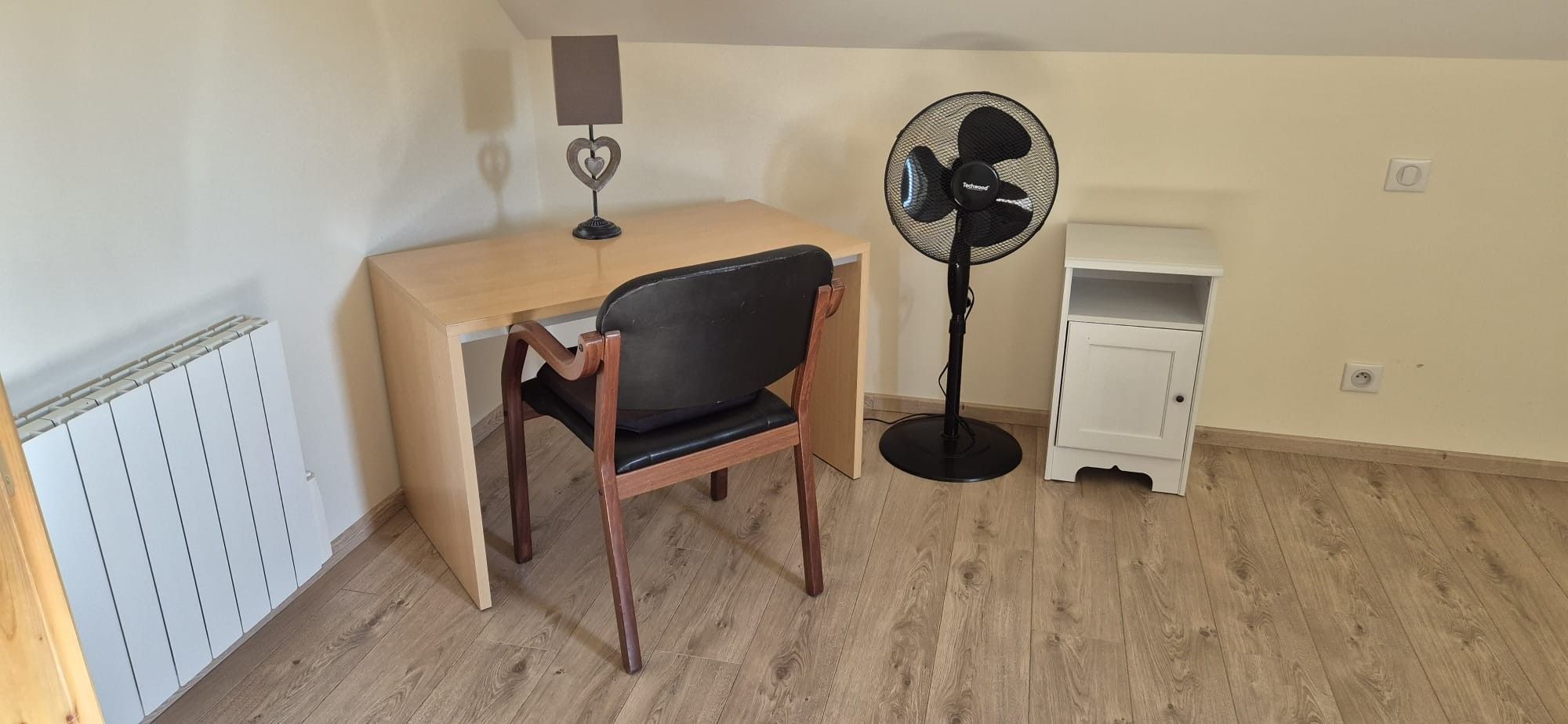 Ein Zimmer mit einem Holztisch, einem schwarzen Stuhl, einem Standventilator, einem kleinen weißen Schrank und einer Heizung.