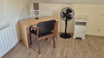 Ein Zimmer mit einem Holztisch, einem schwarzen Stuhl, einem Standventilator, einem kleinen weißen Schrank und einer Heizung.