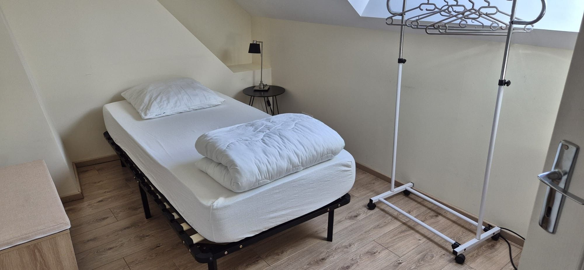 Uma cama de solteiro com colchão e almofadas, uma mesa de cabeceira com abajur e um varal de metal encostado na parede.