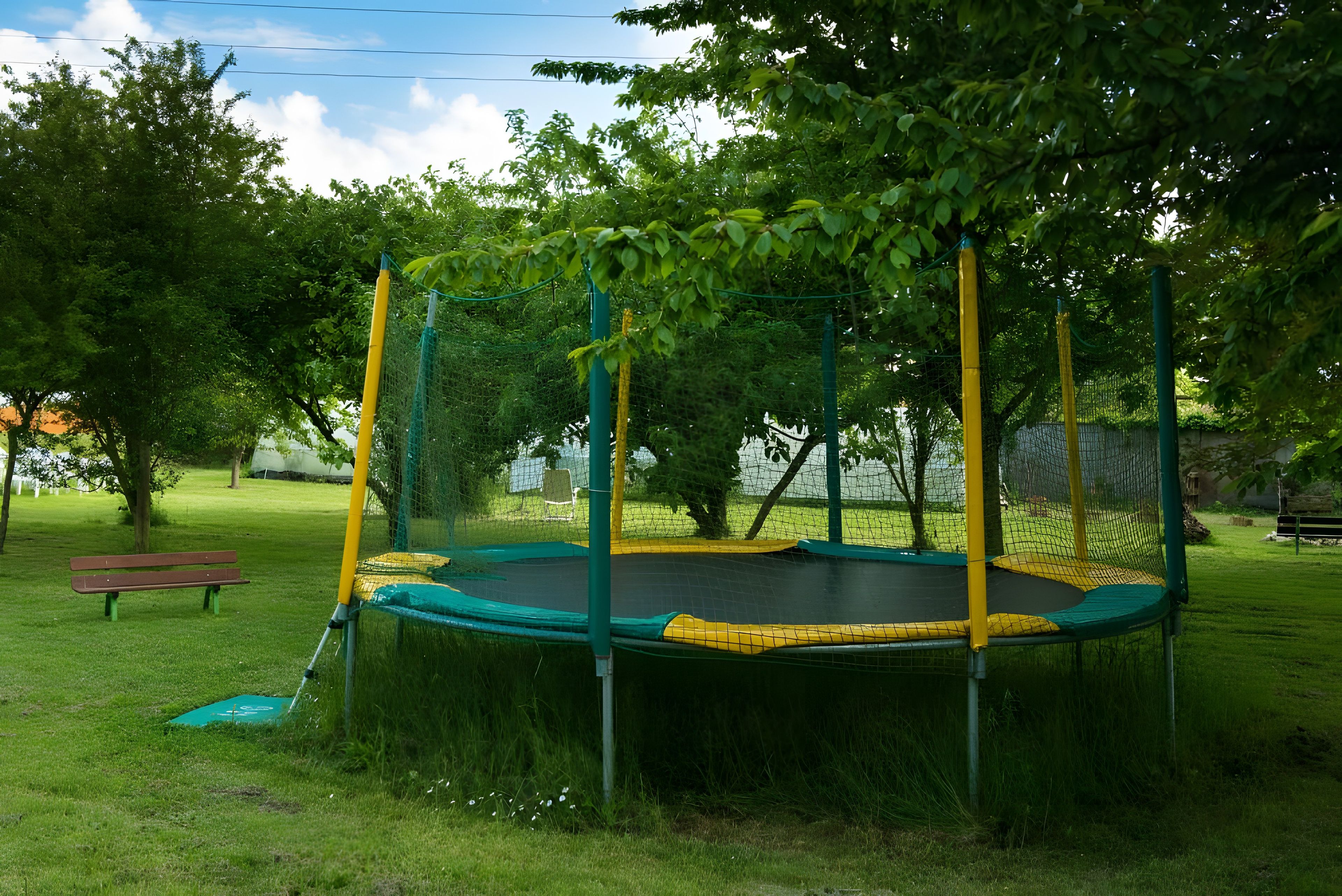Een trampoline omringd door een veiligheidsnet, gelegen in een grasveld met bomen en een bankje op de achtergrond.