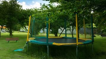 Een trampoline omringd door een veiligheidsnet, gelegen in een grasveld met bomen en een bankje op de achtergrond.