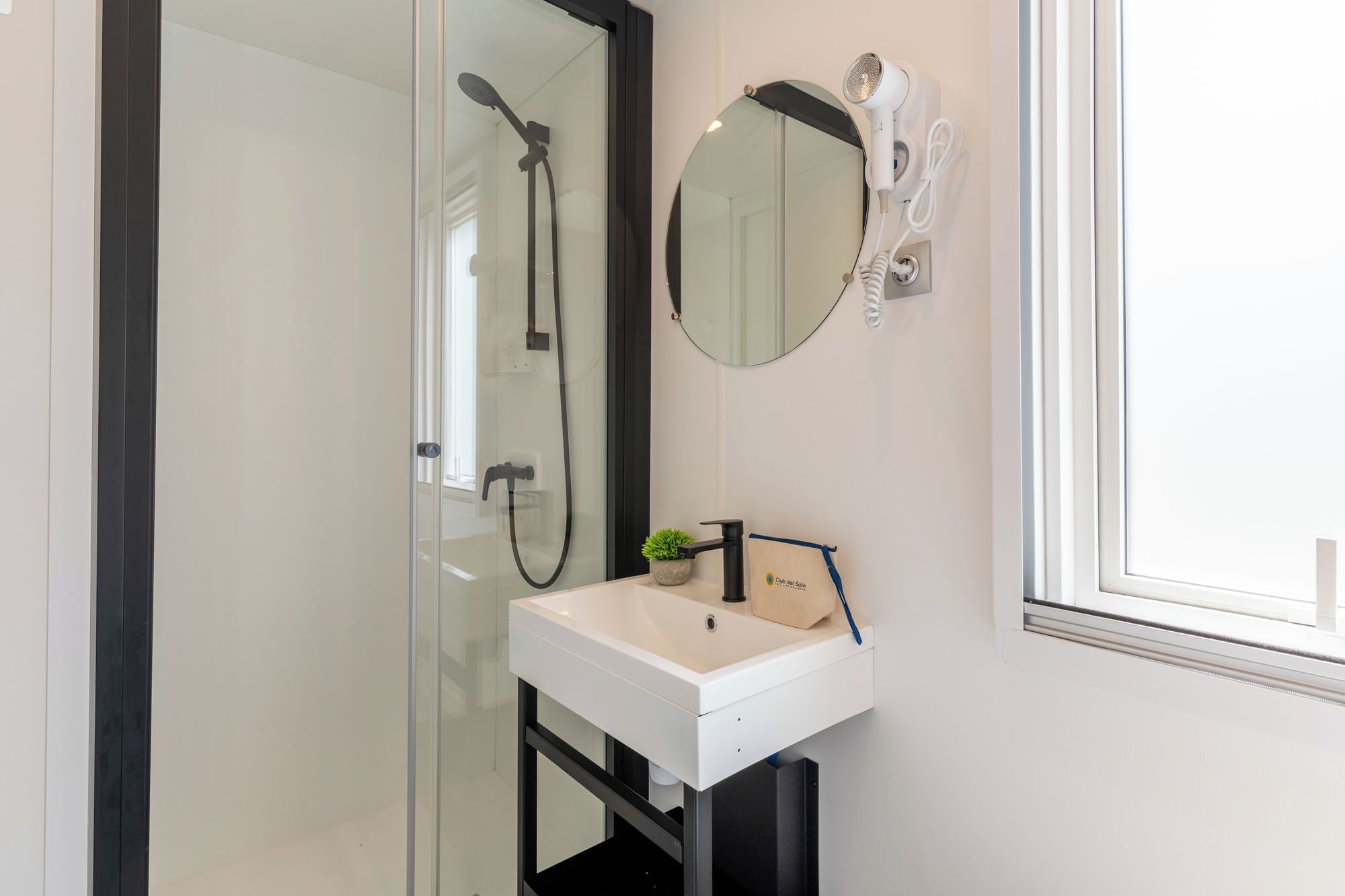 Un baño con cabina de ducha de vidrio, un lavabo blanco, un espejo redondo y un secador de pelo fijado a la pared.