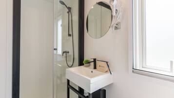 Un baño con cabina de ducha de vidrio, un lavabo blanco, un espejo redondo y un secador de pelo fijado a la pared.