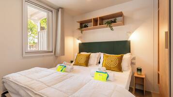 A hotel room with a large bed, a window with curtains, a bedside table, and a shelf with plants.
