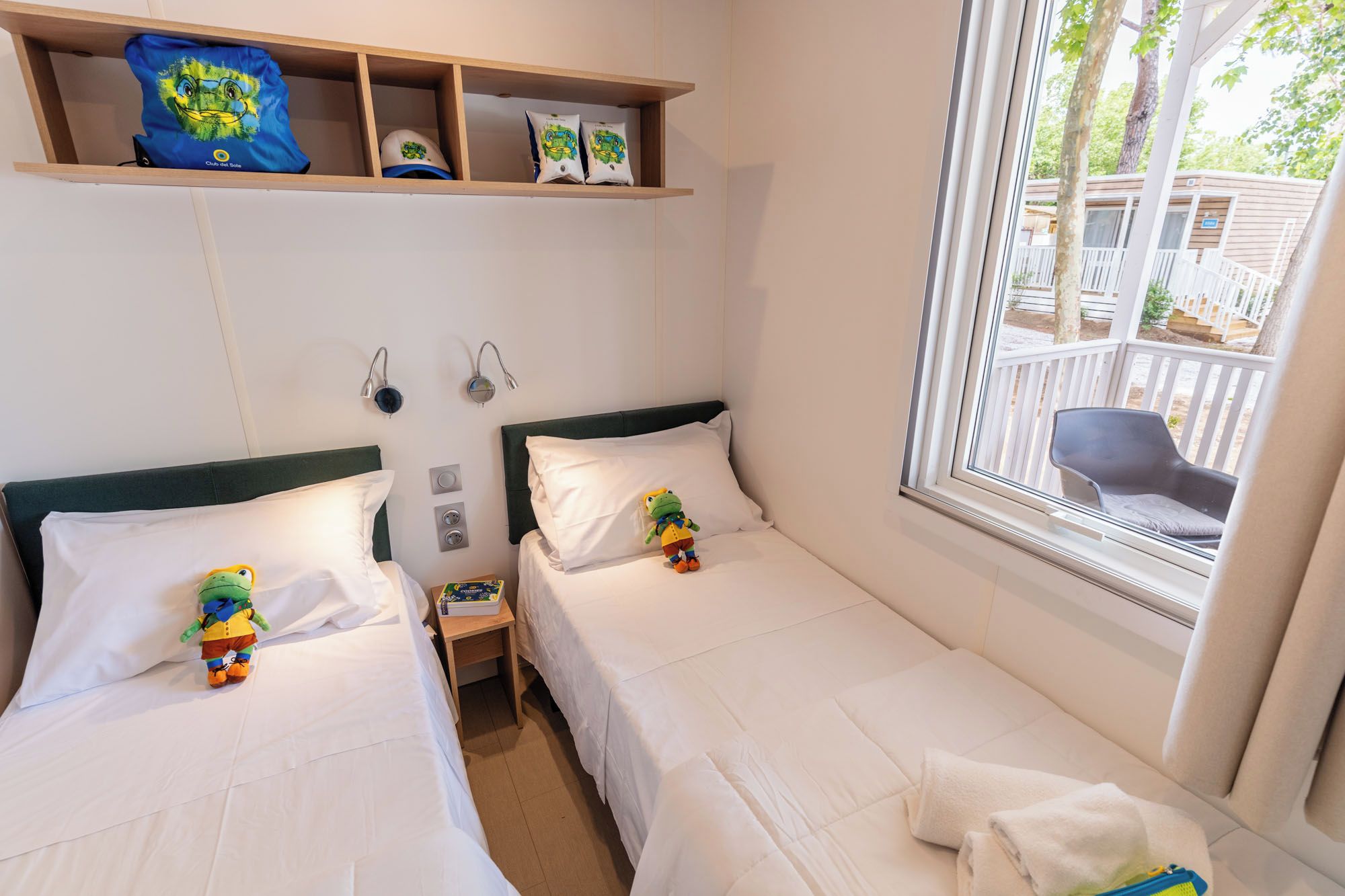 A hotel room with two beds, a shelf with hats, a window with a view, and a small table with a toy.